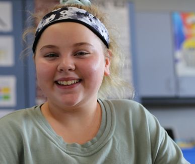 Young student girl, smiling, wearing headband and green shirt, classroom bulletin board in background, Hillsborough Education Foundation
