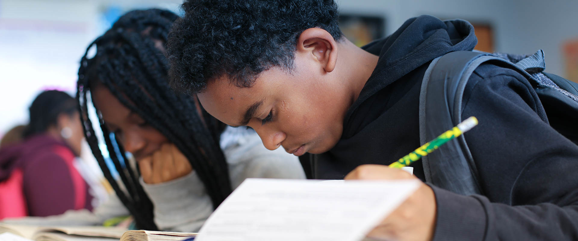 Dos estudiantes, trabajando en el escritorio, con la cabeza gacha, escribiendo en papel con un lápiz en la mano, un niño con capucha negra, una niña con capucha gris, Hillsborough Education Foundation