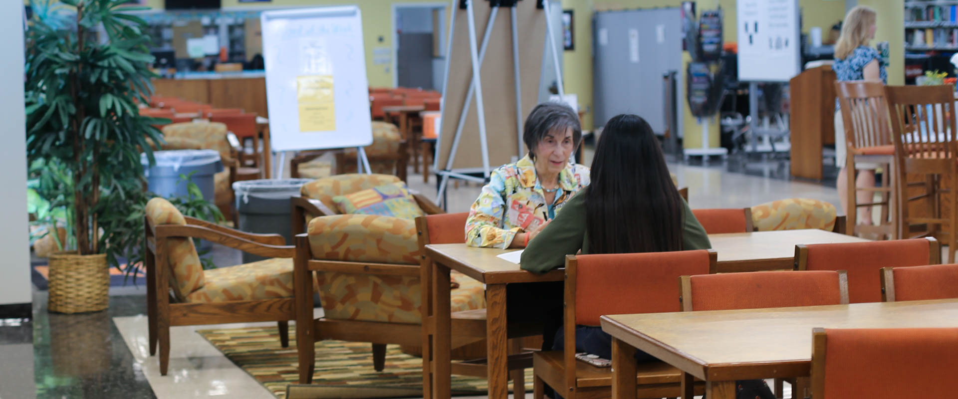 Dos personas sentadas a la mesa en la biblioteca, un mentor y un mentoreado, una mujer mayor hablando con un estudiante más joven de espaldas a la cámara, Hillsborough Education Foundation