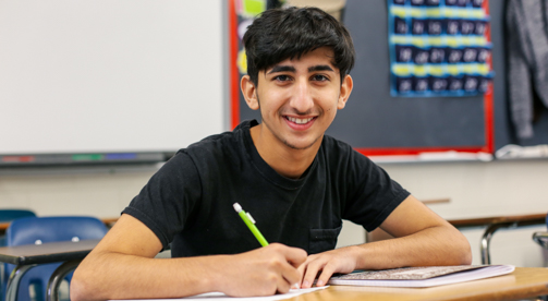 Joven estudiante en el aula con camisa negra, sentado en el escritorio, escribiendo en papel con lápiz en la mano, Hillsborough Education Foundation