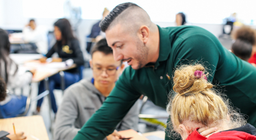 Male teacher in green long-sleeved t-shirt, instructing students at desk, Hillsborough Education Foundation